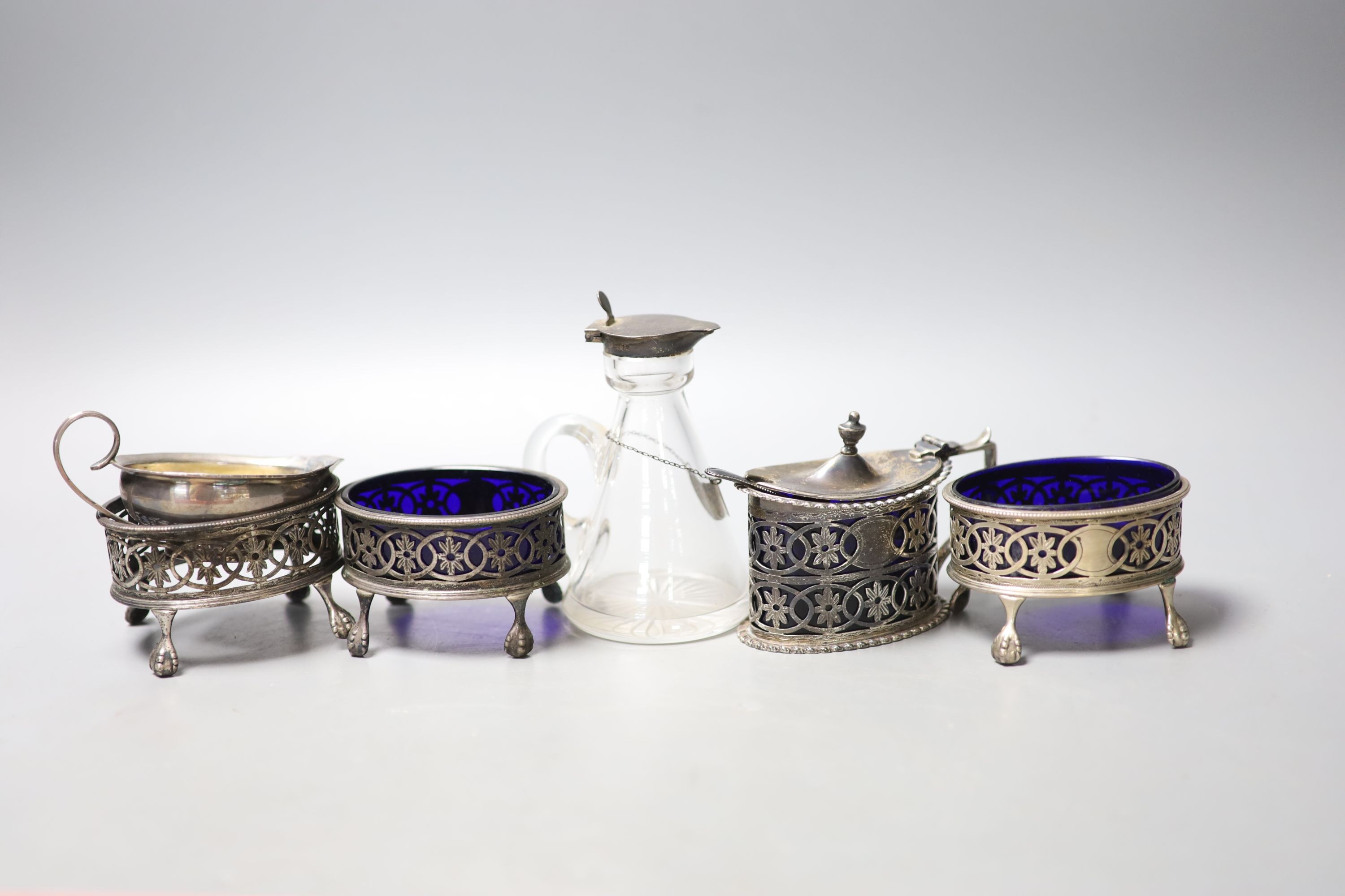 An Edwardian pierced silver oval mustard pot, with liner, a set of three similar salts, one lacking liner, a silver mounted glass whisky tot jug and a small continental jug.